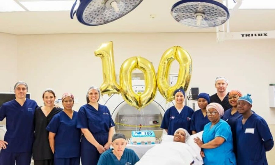 The surgical team celebrating the 100th joint replacement with gold '100' balloons and a cake in theatre.